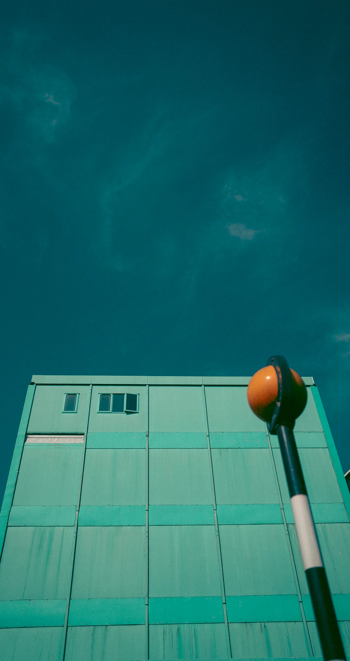 A Belisha beacon leaning across a pale green building under a teal sky